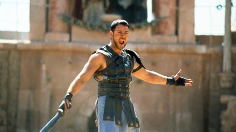 Russell Crowe is dressed like a Gladiator in a leather chest guard and he is wielding a sword. He is shouting and is arms are raised.