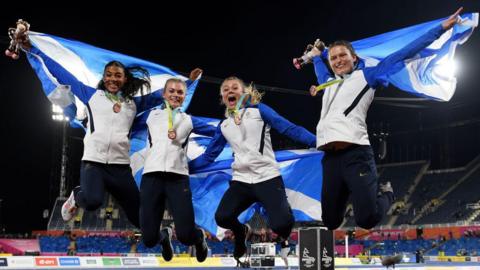 Four women, wearing tracksuits and holding Scottish flags, jump into the air while celebrating winning medals. They are in an athletics stadium.