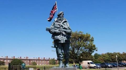 A large bronze statue of a British soldier in the Falklands conflict is is shown with a union jack attached to the back against a clear blue sky. The figure holds a rifle and wears a large jacket.