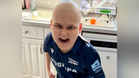 A young boy with a bald head and wearing a dark blue Sales Sharks rugby shirt, does a fierce roaring face at the camera while standing in a kitchen with a sink behind him.