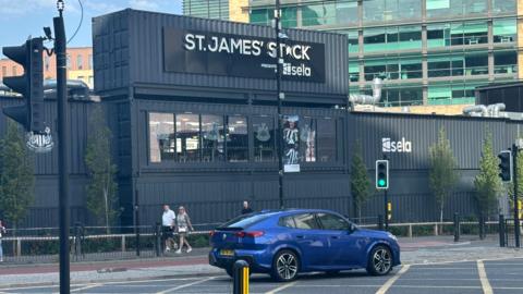 St James' Stack. It is a made up of a number of repurposed shipping containers painted black. A car drives past while a number of people are walking outside. Behind it is a large, tall glass fronted building.