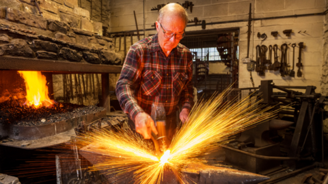 Paul Dennis is an older man with a balding head and a grey fringe of hair. He wears glasses and a checked workman's shirt. He is in his workshop with a lit coal forge to the left of the picture. He is hammering something on an anvil. The photograph is done in long exposure so there are streaks of sparks making a sort of firework effect around the metal being worked. There are lots of metal tools and other machines in the background.