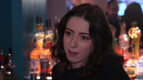 A woman with dark hair is standing inside a pub.