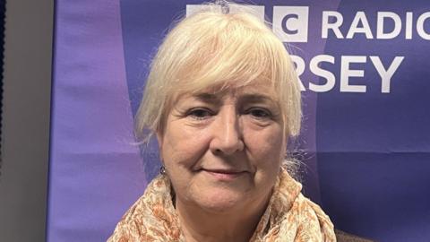 Deputy Karen Wilson is standing in front of a BBC Jersey banner. She is wearing a brown blazer and a floral scarf.