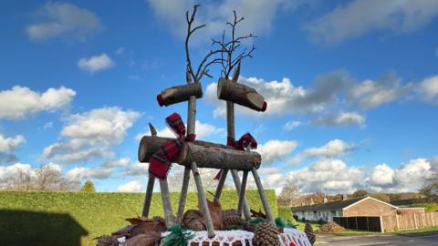 A postbox with reindeer made out of wooden sticks and logs surrounded by pine cones. The postbox is on a residential street. The sky is blue and there's a green clipped hedge behind.
