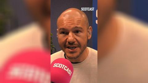 A man is sitting in a studio, speaking into a red mic that reads 'Scotcast'