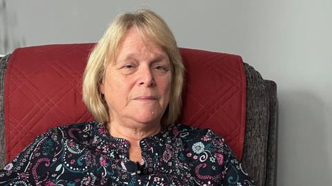 A woman in her sixties is sitting in an armchair with a serious expression on her face. She is dressed in a black blouse with pink swirls and has light blonde hair.