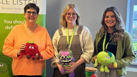 Sarah Kelly from Manx Lottery Trust and Isle Listen's Becks Macnair and Holly Ramsay stand behind the table smiling for the camera. They are holding stuffing children's toys used in the lessons.