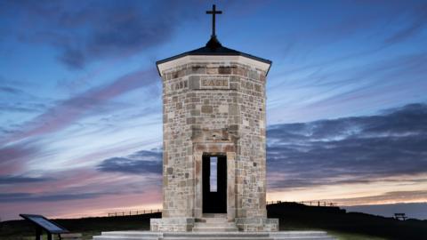The Compass Point tower is lit up at twilight with a blue, pink, purple and orange sky behind it. The stone octagonal tower has a cross on the top of it and the lettering EAST above an open doorway which shows a narrow window without glass beyond. There is a visitors' guide board to the left of it.
