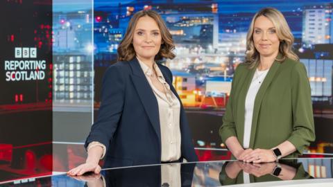 Laura Miller and Laura Goodwin standing behind the Reporting Scotland desk