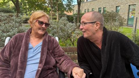 Woman in purple dressing gown and light blue top. She is wearing sunglasses and holding hands with a man in a black dressing gown looking back at her.