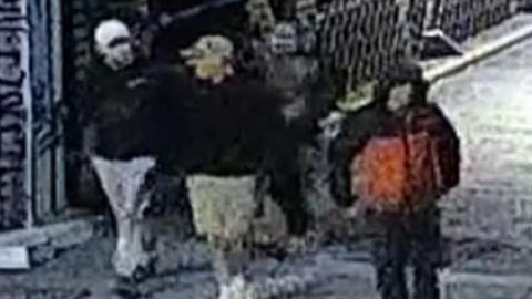 A blurry CCTV image of four men wearing jackets and baseball caps in a street at night time.