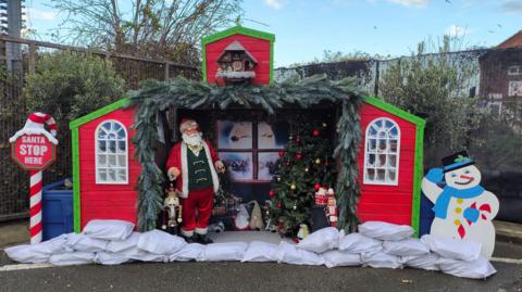 A red house with green trim houses Father Christmas and a Christmas window with a reindeer and sleigh flying. On the left is a Santa stop here sign and on the right a snowman doing a salute.