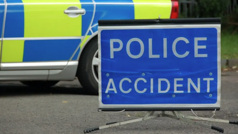 A blue police accident sign in front of the back wheel of a police car.