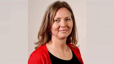 Amanda Clare has long brown hair and is pictured wearing a black top and red cardigan; she is smiling at the camera.