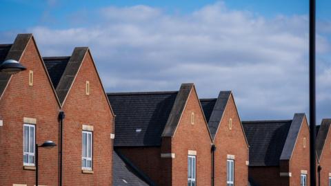 The image shows a new build house development estate; the bricks are red and brown and all the houses have one window facing outward. The shy is cloudy and slightly grey.