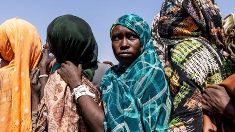 The heads and shoulders of three women standing close together in a queue. They are wearing headscarves - one orange, one black and one turquoise and two are facing away from the camera and one is looking towards it.