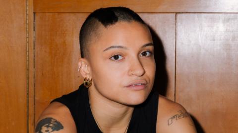 Rene Matić. They have short black hair and are wearing a black vest top. They are sitting in front of a wooden wall.