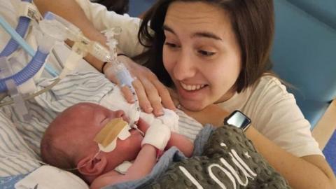 A woman sat beside a bed smiling at a sleeping baby