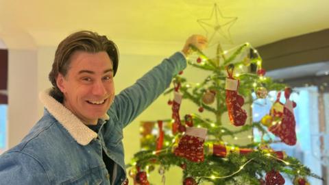 A dark haired man, wearing a denim jacket, is holding a star at the top of a decorated Christmas tree