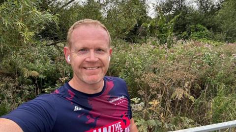 A man in a blue and red sports t-shirt smiles at the camera as he takes a selfie. He has white airpods in his ears and stands in front of some bushes.