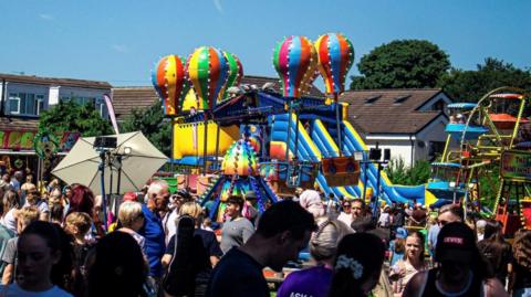 A lively outdoor carnival scene on a sunny day, featuring colourful amusement rides such as hot air balloon-themed attractions, inflatable slides, and a Ferris wheel. The area is crowded with people enjoying the event, surrounded by vibrant stalls and signage.