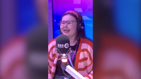 A young girl wearing a brightly coloured cardigan and headband smiling and speaking into a BBC Radio 4 mic in a radio studio