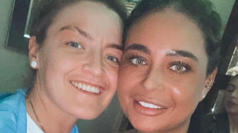 A woman with dark blonde hair and blue eyes is wearing a white top, a blue jacket and chain around her neck. She is next to another woman with black hair and hazel eyes.