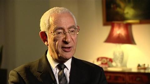 Lord David Triesman, wearing a black suit and speaking to the camera, in the dimmed light coming from a red lampshade