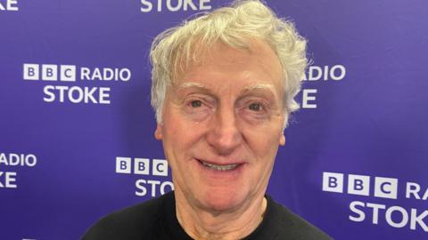 A man with white hair wearing a black t-shirt pictured against a purple backdrop for Radio Stoke. He is smiling.