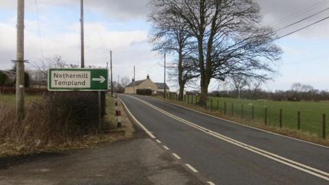 The A701 in Dumfries and Galloway with trees and a fence at the side and a sign for Nethermill and Templand