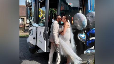 Danny and his wife are standing by the bin lorry, kissing. Balloons are next to them. The bin lorry door is open. The lorry is parked on a residential street.