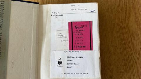 An open book with the words 'Read, G, Music notation' written. There is a list of dates which date back to 1973 of when it was taken out. It also says 'Cornwall County Library, County Hall, Truro. Please return books promptly'.