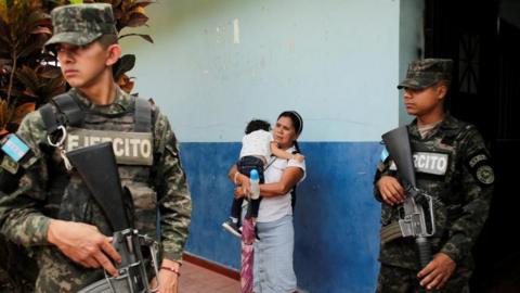 Members of security forces stand guard as a woman carries her daughter while waiting outside a polling station, as counting continues in San Antonio de Flores, Honduras, on 7 December, 2025.