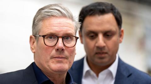 Sir Keir Starmer in the foreground. He has grey hair and dark-rimmed glasses and is looking straight at the camera. Only his head and shoulders are visible. Anas Sarwar is in the background. He is wearing a blue suit over a white shirt and has dark hair.