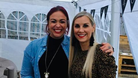 Lois Sherret smiling to camera, wearing a black top, blue shirt and putting her arm around Claire Berridge who has long, blonde hair and is wearing a leopard print top