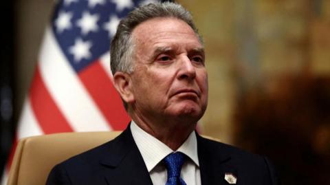 Steve Witkoff wearing a suit and blue tie sits in front of a US flag