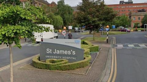 The entrance to St James's University Hospital, indicated by a sign on a traffic island. The traffic island also has plants and trees. Zebra crossings are present on the road either side of the island and a large redbrick building can be seen in the far background.