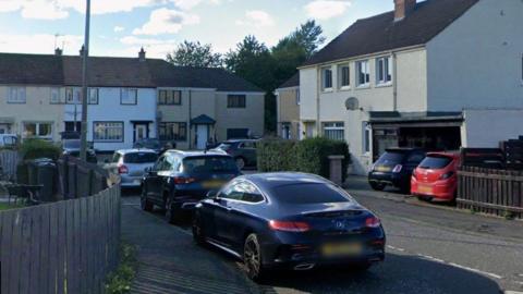 A cul-de-sac with brown and white houses. There are trees behind the houses at the far end. There are three cars in the foreground.