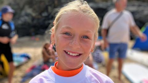 Ellawyn, who is a young girl with blue eyes and blonde hair tied back, smiling at the camera on the beach.