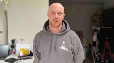 Joe Bishop, who served in the RAF, is pictured wearing a dark grey hoodie with the Building Bridges logo. He is a bald white man with hints of a ginger beard