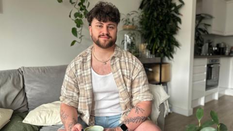 A man with dark brown hair and a beard sits on a sofa smiling at the camera. He's wearing a checked beige shirt over a white vest and has tattoos