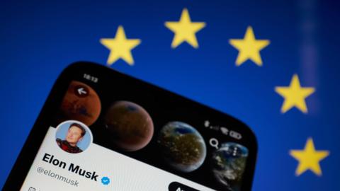 A picture of a phone against the backdrop of the blue and yellow EU flag with yellow stars. The phone has Elon Musk's X profile on it with his face and a blue tick next to it.
