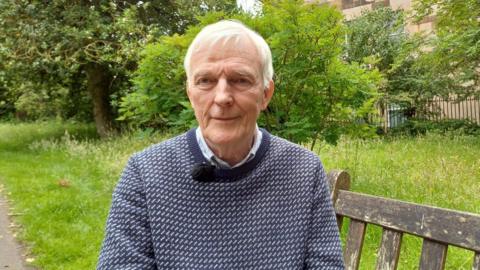 Coinneach MacIlleathain na shuidhe air beinge sa ghàrradh agus coltas gu math toilichte air.