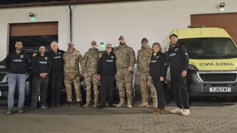 Four men in Khaki army uniforms stand with six people in black clothes. An ambulance is parked to the side of them.