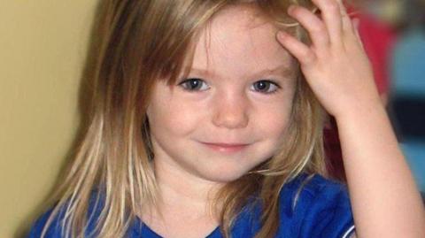 Madeleine McCann, aged three, looks into the camera as she wears a blue and white football top. Her left hand is raised and brushing against her hair.