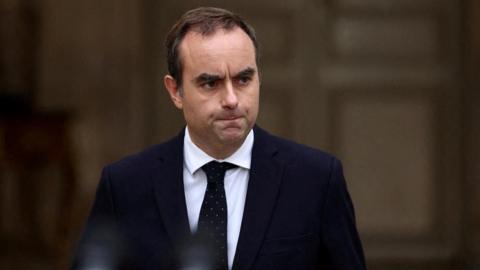 French Prime Minister Sebastien Lecornu arrives to deliver a statement at the Hotel Matignon in Paris. He is wearing a dark suit with a white shirt and dark-coloured tie with small spots on it. He stands in front of a brown background. He has a stern expression.