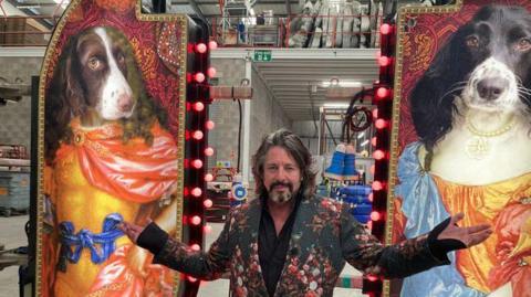 Laurence Llewelyn-Bowen, dressed in a multi-patterned green jacket and black shirt, has long brown hair and a beard with grey tufts. He is standing either side of images of two spaniels wearing royal costumes.