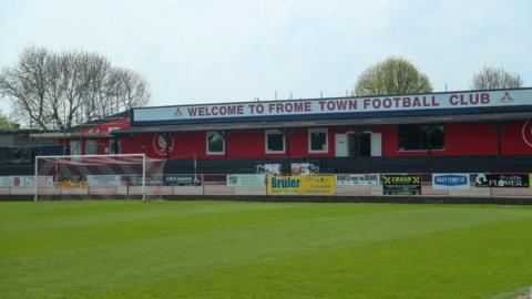 A football pitch with the club building behind it. The building is painted red and says Welcome to Frome Town Football Club on the roof.