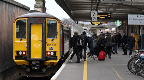 A train at Shrewsbury station with passengers disembarking from the right hand side of the train onto the platform with a sign above saying 7b. Some bikes stored in a rack can be seen on the right of the image.